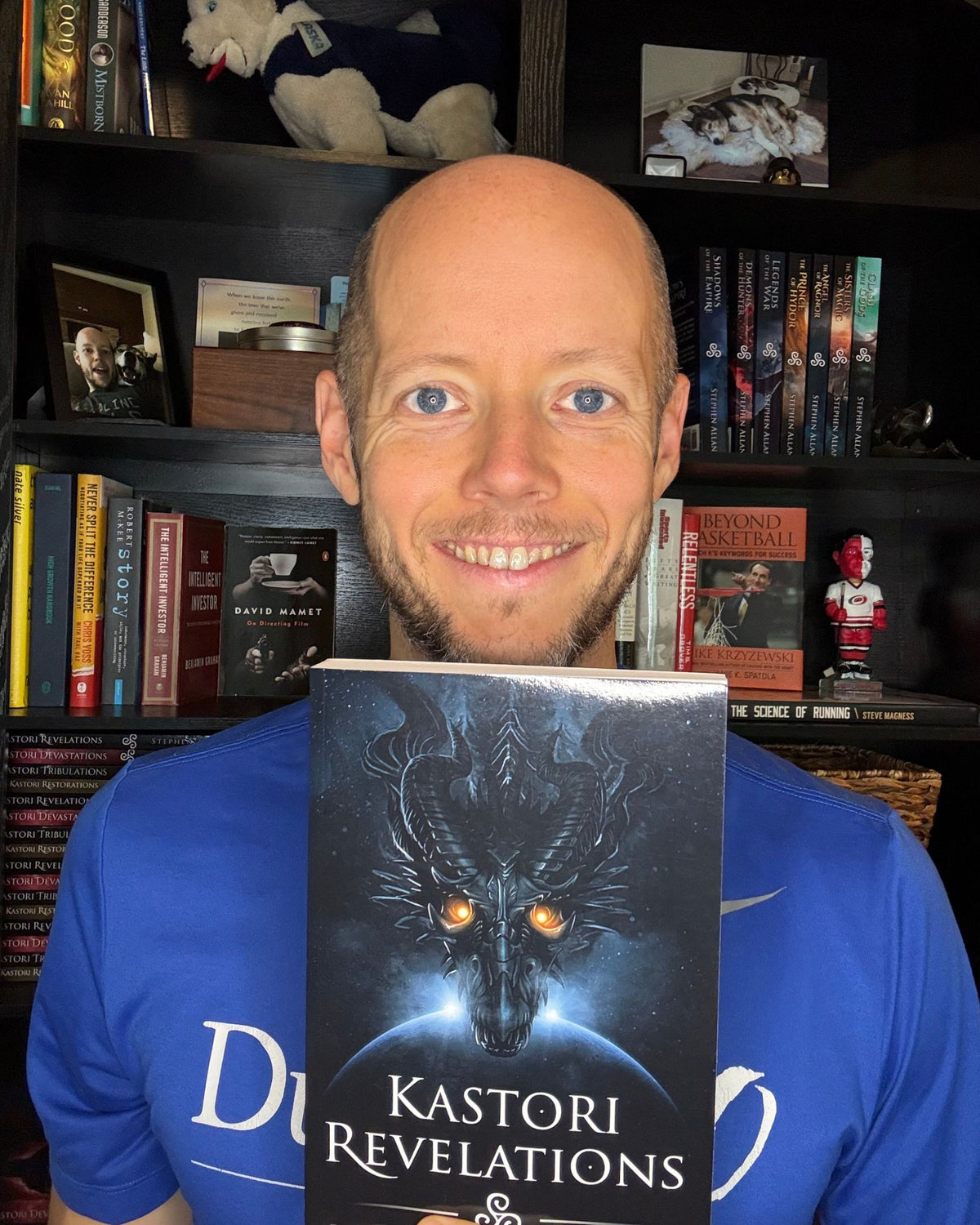 Author Stephen Allan holding a paperback copy of Kastori Revelations in front of his bookshelf
